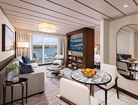 The Grand Suite on American Patriot, with large floor-to-ceiling sliding glass doors leading to a private balcony overlooking the New England coast. There is a grey sofa facing a large wooden TV stand in the center of the room, with a TV and shelves with decor. 