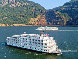 Two river cruise ships, including a classic paddlewheel boat and a modern riverboat, sailing past each other along the Columbia River Gorge.