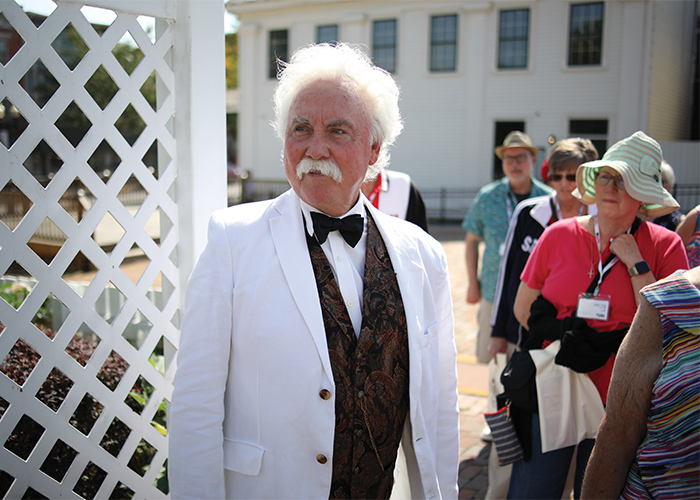 A distinguished older man impersonating Mark Twain with white hair and a white mustache, wearing a white suit and bow tie, showcasing a refined and classy appearance.