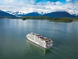 An aerial view of a multi-deck cruise ship navigating a wide bay with snow-capped mountains and evergreen forests lining the shore during an Alaska cruise.