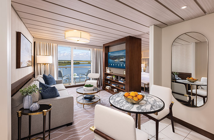 The Grand Suite on American Patriot, with large floor-to-ceiling sliding glass doors leading to a private balcony overlooking the New England coast. There is a grey sofa facing a large wooden TV stand in the center of the room, with a TV and shelves with decor. 