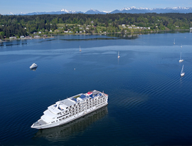 The cruise ship American Constellation travels across a wide, sunlit bay dotted with several sailboats, with a forested coastline and distant mountain range in the background.