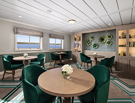 The game room on a ship in the Puget Sound featuring deep green velvet armchairs, round wooden tables, and decorative green wall accents, with large windows overlooking a scenic water view.