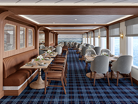 The restaurant on American Encore, where there are several round tables and navy blue chairs along the blue and white patterned carpet, and two round chandeliers hanging from the ceiling. There are large windows with clear views of the Columbia River on one wall, and floor-to-ceiling paintings on the back wall.