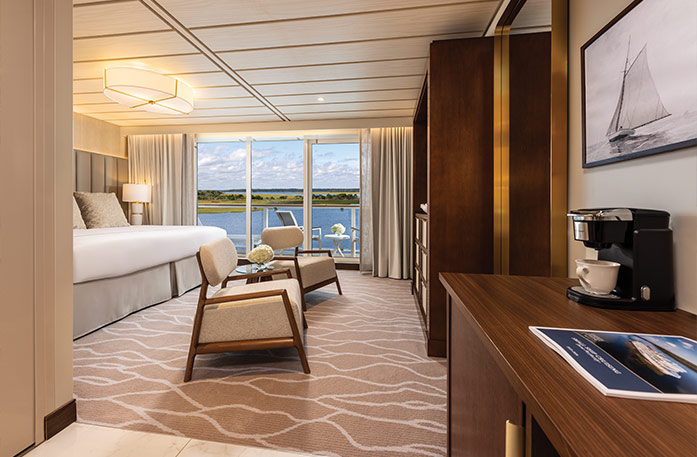 An elegant Sky Suite aboard an American Cruise Lines ship, showcasing a spacious layout with premium wood cabinetry, a polished seating area, and a brightly lit bedroom.