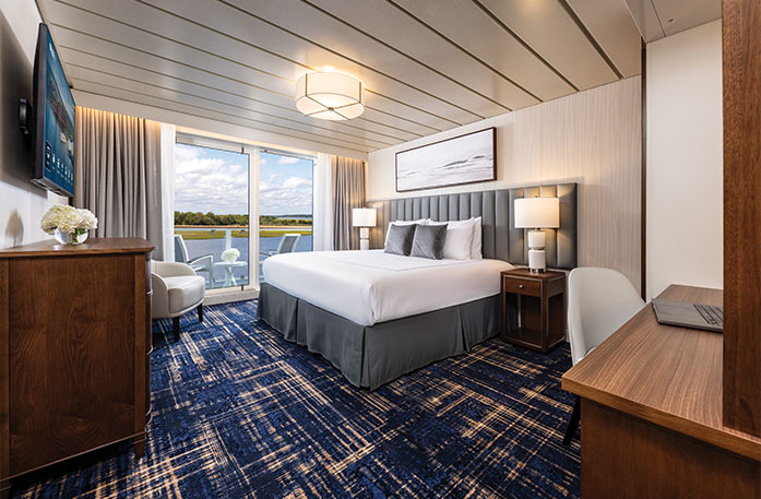 The Vista Balcony stateroom on American Patriot, with floor-to-ceiling sliding glass doors that lead to a private balcony overlooking the New England coast. There is a bed against the wall on the right side of the room and an end table with a lamp on each side.