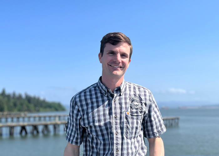 Man in American Cruise Lines shirt stands by the water with a wooden pier and trees in background.