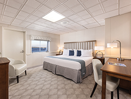 The Deluxe Balcony 2 stateroom on American Spirit, an American Cruise Lines small ship with a push door leading to a private balcony, beside a large window overlooking the Chesapeake Bay. There is a bed on the right side of the room with an end table with a lamp on top on each side of the bed. Across from the bed, there is a cream-colored chair  beside a wooden dresser, with a TV on the wall.