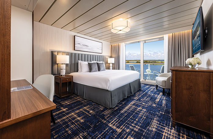 The Premium Balcony stateroom on American Pioneer, with floor-to-ceiling sliding glass doors that lead to a private balcony overlooking the Florida Gulf Coast. There is a bed against the wall on the left side of the room and a TV on the wall.