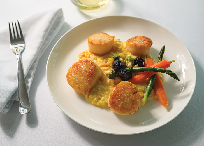 A plate with four scallops and vegetables on top of risotto served on American Cruise Lines.