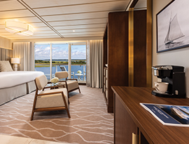 A modern cruise ship suite featuring a bed, seating area with two armchairs, and large glass doors opening to a private balcony with water views.