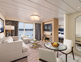An expansive luxury cruise ship suite featuring a modern living area with a sofa, glass coffee table, and balcony access, separated from the bedroom by a TV wall divider, with a small dining table in the foreground.
