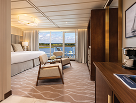 An elegant stateroom on the American Pioneer, featuring two armchairs, a large bed, and a private balcony with sweeping views of the water.