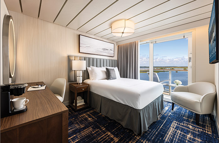 The Single Balcony stateroom on American Patriot, with floor-to-ceiling sliding glass doors that lead to a private balcony overlooking the New England coast. There is a bed against the wall on the left side of the room and a TV on the wall.