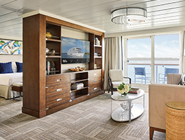A spacious luxury suite on a modern river cruise ship, featuring a private living area with elegant wood furnishings and sliding glass doors that open to a balcony with outdoor seating overlooking the river.