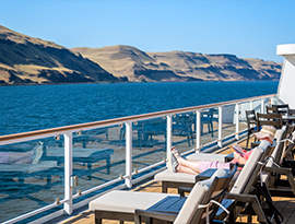 Guests lounging on the outdoor observation deck of a riverboat as it cruises past the expansive, sunlit hills of the Columbia and Snake River region.