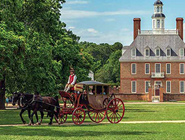 A traditional horse-drawn carriage traveling across a scenic green lawn in front of the historic Governor's Palace in Colonial Williamsburg, with dense trees flanking the brick estate under a clear sky.