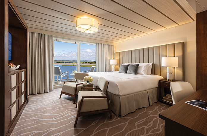 The Veranda Suite on American Patriot, with floor-to-ceiling sliding glass doors that lead to a private balcony overlooking the New England coast. There is a bed against the wall on the right side of the room with blue accent pillows and large TV stand across from it.