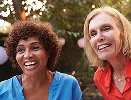 Two women, one in a blue shirt and the other in a red shirt, are smiling and leaning forward in a garden with string lights in the background. 