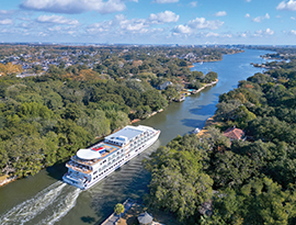 An aerial view of a modern multi-deck cruise ship navigating a narrow coastal river lined with dense green trees and residential properties under a clear sky.