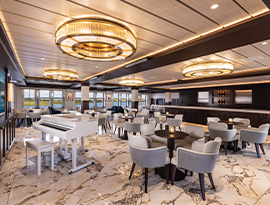 The elegant lounge on the American Pioneer, featuring a white grand piano, polished marble flooring, and circular pendant lighting, with comfortable seating overlooking the scenic water views.