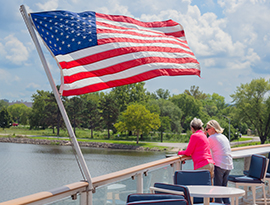 Two passengers standing at the railing of an outdoor ship deck, looking out at the green shoreline of the Mississippi River next to a large American flag waving in the breeze.