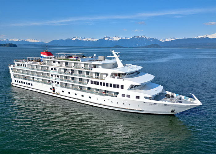 A white Constellation Class American Cruise Lines ship sails on calm waters with snow-capped mountains in the background.