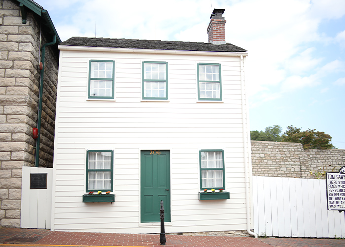 Mark Twain's light cream colored house with a dark green door and window planters in Hannibal, to the left of the whitewashed fence of Tom Sawyer.