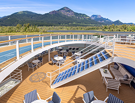 A multi-level outdoor deck on American Encore featuring outdoor seating, a walking track, and stairs, looking out toward a wide river and forested mountains under a clear sky.
