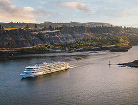 An aerial view of an American Riverboat cruising through the Columbia River Gorge, where there is a small island formation in the middle of the water and mountains surrounding