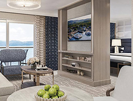 The Grand Suite on American Patriot, with large floor-to-ceiling sliding glass doors leading to a private balcony overlooking the New England coast. There is a tan sofa facing a large wooden TV stand in the center of the room, with a TV and shelves with decor. 