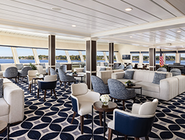 A spacious ship lounge featuring modern sofas, patterned carpeting, and large windows offering panoramic views of the Alaskan shoreline.