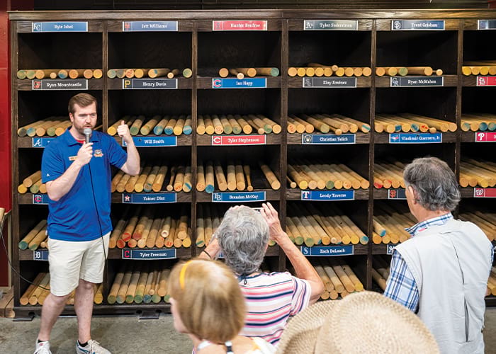 A wall with several bins each containing a variety of wooden baseball bats, with a name label above each shelf at the Louisville Slugger Museum in Louisville. There is a man with a microphone presenting to American Cruise Lines guests.