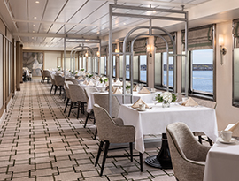 An elegant cruise ship dining room featuring tables set with white linens and floral centerpieces, lined with large windows overlooking the water.